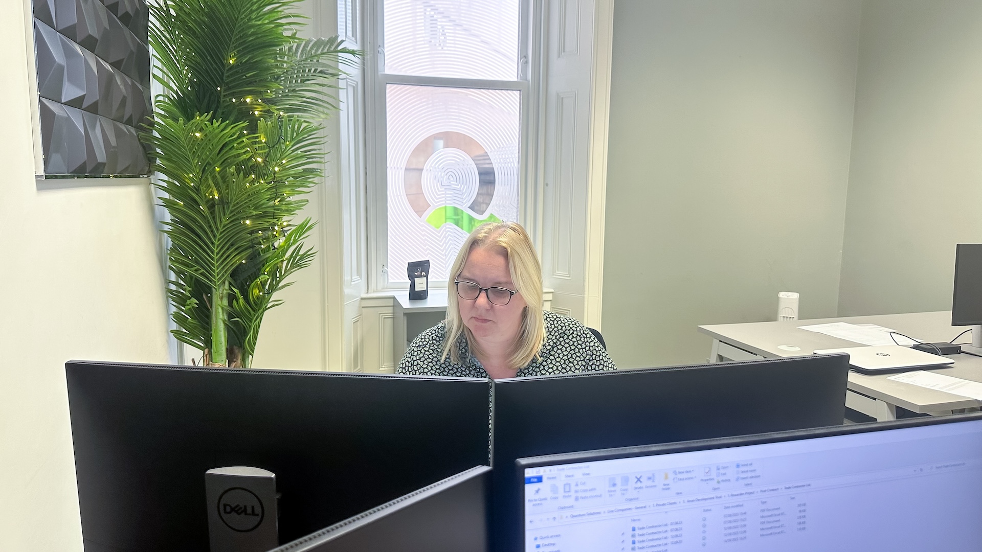 Blonde haired woman looking at two screens with a green plant and window in the background